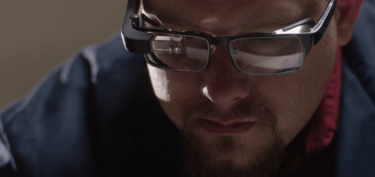 A close-up of a person wearing smart glasses with reflective lenses. They have a short beard and are concentrating intently, with shadows adding depth to their face. The glasses feature thick, dark frames, reminiscent of a design popular in the enterprise market, and the person is dressed in a dark-colored outfit.