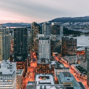 Aerial view of a modern cityscape in Canada at dusk, with numerous high-rise buildings illuminated by streetlights. In the background, a calm body of water and mountainous terrain are visible under an overcast sky. Apple Maps vehicles can be seen expanding their coverage through the city's streets.