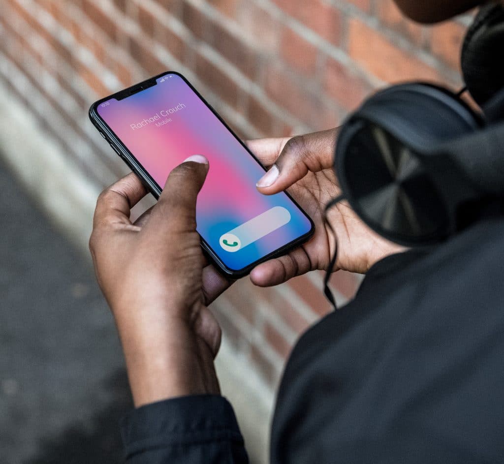 A person, wearing headphones, holds an Apple smartphone displaying an incoming call from "Richard Couch." The phone shows the option to accept the call using Haptic Touch. The background includes a brick wall and part of a street.