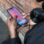 A person, wearing headphones, holds an Apple smartphone displaying an incoming call from "Richard Couch." The phone shows the option to accept the call using Haptic Touch. The background includes a brick wall and part of a street.