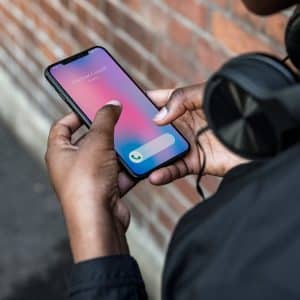 A person, wearing headphones, holds an Apple smartphone displaying an incoming call from "Richard Couch." The phone shows the option to accept the call using Haptic Touch. The background includes a brick wall and part of a street.