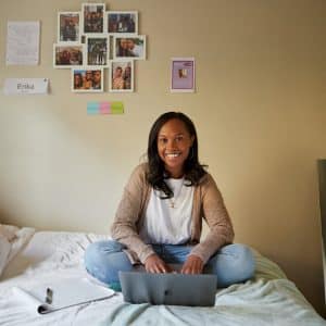 A young woman sits cross-legged on a bed with a laptop in her lap, smiling at the camera. She is in a bedroom with photos, notes, and a name tag "Erika" attached to the wall behind her. An Apple notebook and pen are beside her on the bed, reflecting the spirit of diversity celebrated at WWDC.