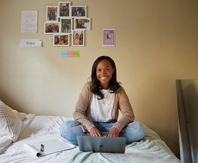 A young woman sits cross-legged on a bed with a laptop in her lap, smiling at the camera. She is in a bedroom with photos, notes, and a name tag "Erika" attached to the wall behind her. An Apple notebook and pen are beside her on the bed, reflecting the spirit of diversity celebrated at WWDC.