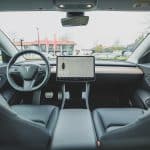 Interior view of a modern electric car, reminiscent of Apple's design ethos, featuring a minimalist dashboard with a large central touchscreen display, steering wheel with a logo, and sleek black seats. The windshield shows an outdoor scene with some buildings and trees in the background.