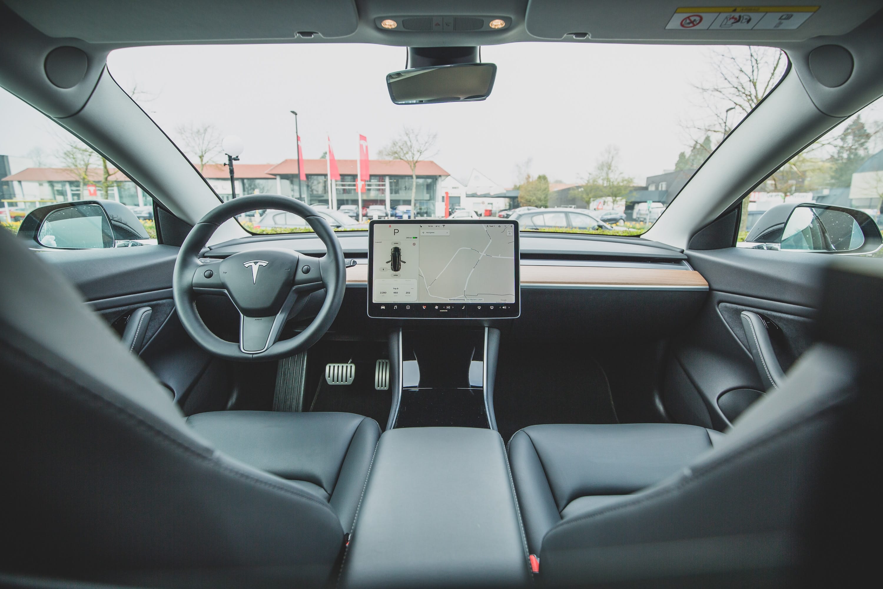 Interior view of a modern electric car, reminiscent of Apple's design ethos, featuring a minimalist dashboard with a large central touchscreen display, steering wheel with a logo, and sleek black seats. The windshield shows an outdoor scene with some buildings and trees in the background.