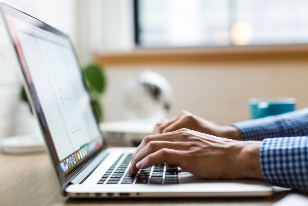 A person with a blue checkered shirt types on a laptop placed on a wooden desk. There is a green plant and a blue coffee mug on the desk. The background shows a blurred window and some indistinct objects, perhaps open tabs in Safari, reflecting Apple's clean design while browsing.