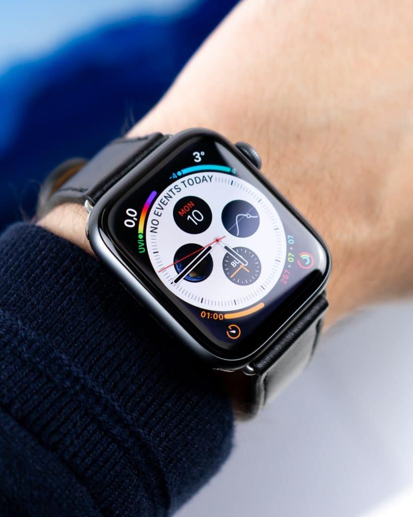 Close-up of a person's wrist wearing an Apple Watch 5 with a black band. The watch display showcases various Apple Watch concepts, including colorful activity rings, date, time, weather, a heart rate graph, and multiple widgets. The background is blurred blue and white.