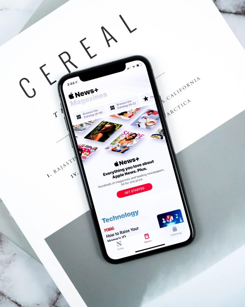 A smartphone displaying the Apple News+ app screen is placed on a stack of magazines, with the top magazine titled "Cereal". The app screen showcases various magazine covers and the headline "Everything you love about Apple News, plus hundreds of magazines at your fingertips," highlighting Apple's offerings since Q1 2019.