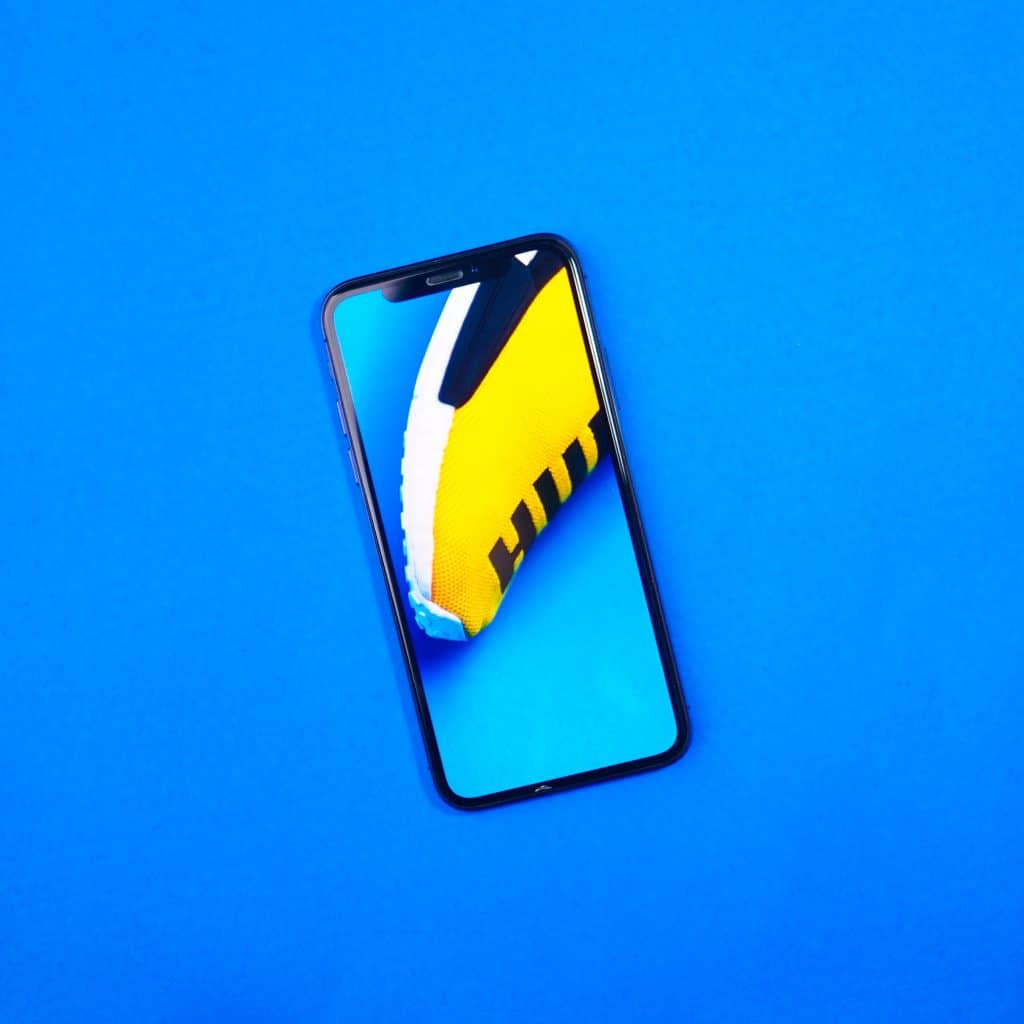 A 2019 iPhone with a notch display is lying on a bright blue surface. The screen shows a close-up image of a yellow and white shoe with black stripes against the same blue background. The phone is oriented at a slight angle.