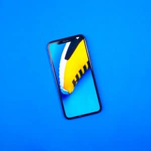 A 2019 iPhone with a notch display is lying on a bright blue surface. The screen shows a close-up image of a yellow and white shoe with black stripes against the same blue background. The phone is oriented at a slight angle.