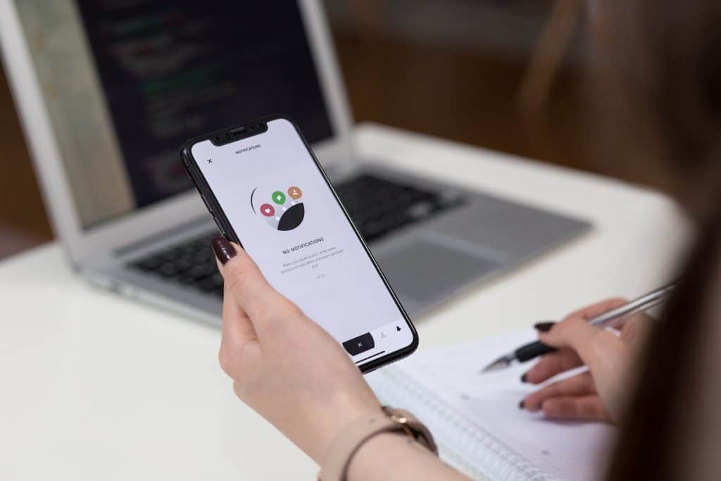 Person holding a smartphone with an iOS productivity app displayed, near an open laptop with visible code. They are taking notes in a notebook with a pen, sitting at a white desk, ready to get more done. The background is slightly blurred.