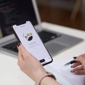 Person holding a smartphone with an iOS productivity app displayed, near an open laptop with visible code. They are taking notes in a notebook with a pen, sitting at a white desk, ready to get more done. The background is slightly blurred.