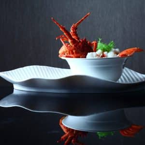 A gourmet dish featuring a cooked lobster, garnished with fresh greens, presented in a sleek, white bowl placed on an elegantly curved, white porcelain plate. With a dark, reflective background enhancing the fine dining presentation, this creation embodies FoodTech innovation.