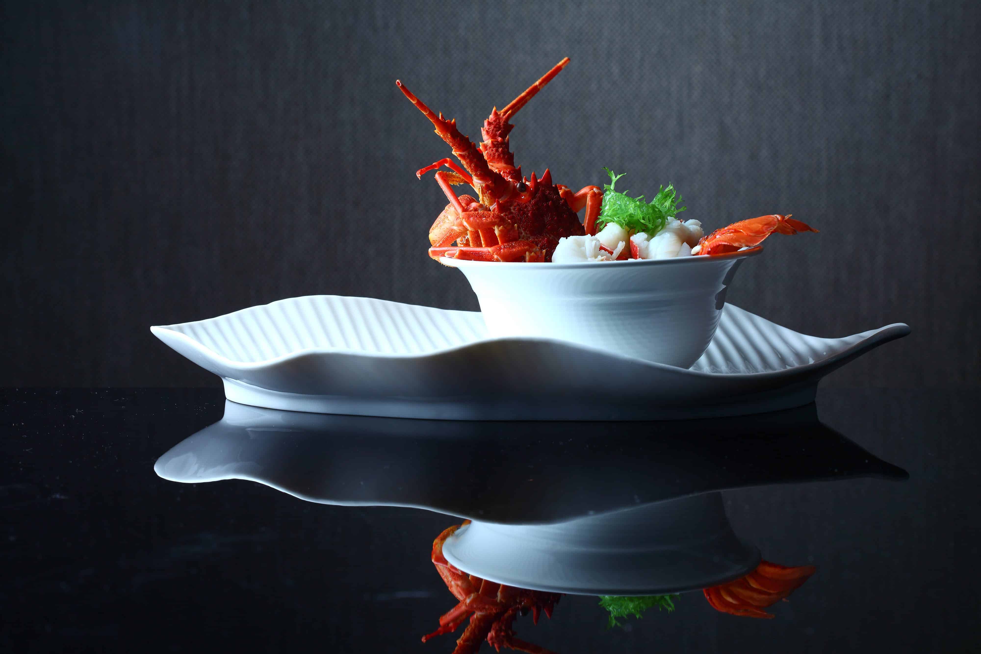 A gourmet dish featuring a cooked lobster, garnished with fresh greens, presented in a sleek, white bowl placed on an elegantly curved, white porcelain plate. With a dark, reflective background enhancing the fine dining presentation, this creation embodies FoodTech innovation.