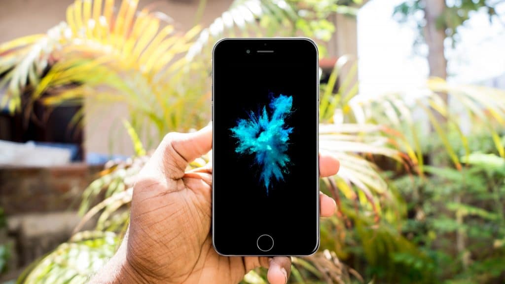 A hand holding an iPhone 6 with a black screen displaying a vibrant blue and green abstract splash. The background features lush, green foliage and a sunny outdoor setting, perfectly capturing the essence of a bright day.