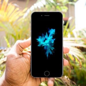 A hand holding an iPhone 6 with a black screen displaying a vibrant blue and green abstract splash. The background features lush, green foliage and a sunny outdoor setting, perfectly capturing the essence of a bright day.