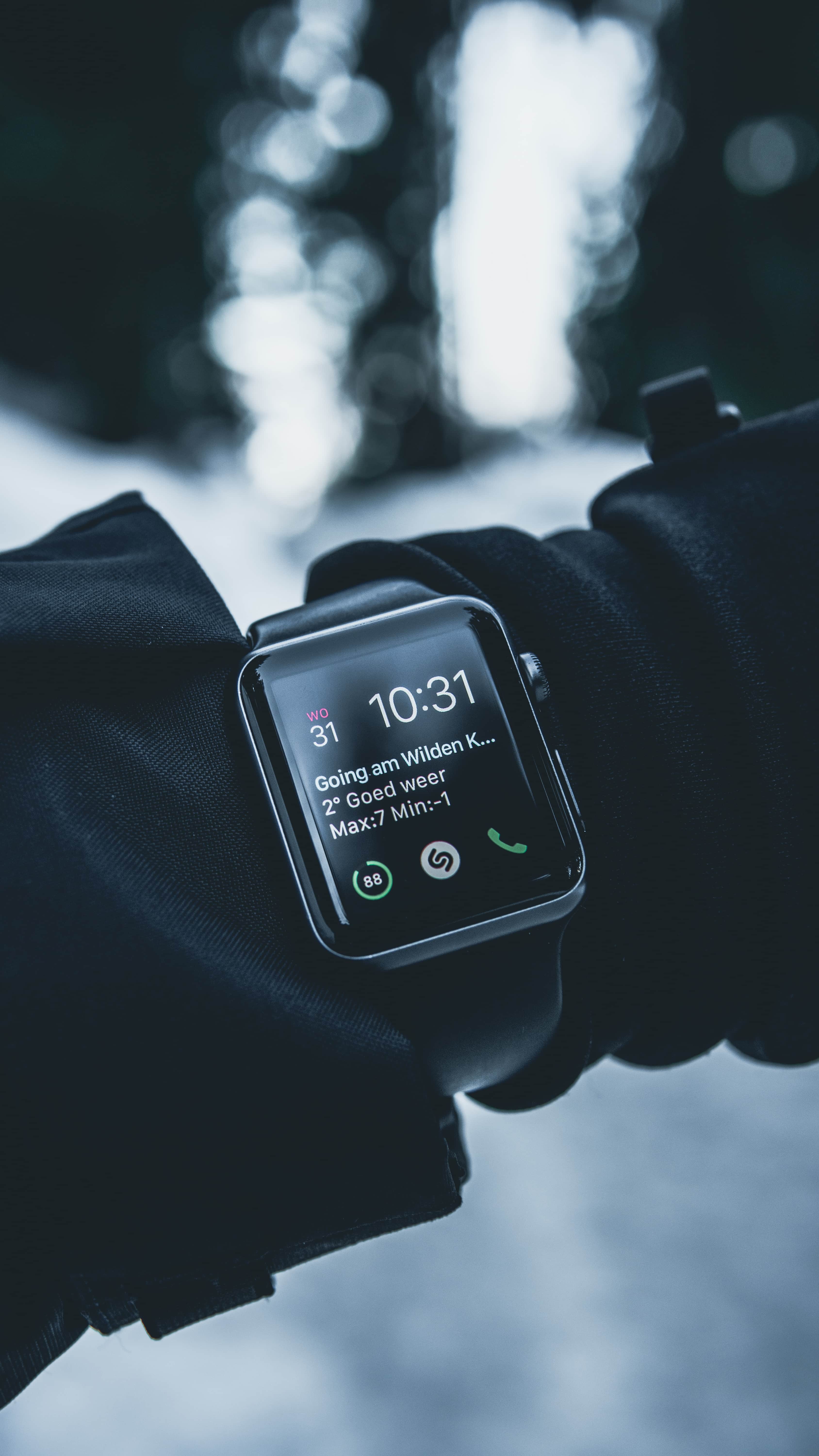 A person wearing a black glove and an Apple Watch, displaying the time as 10:31 and a message about the weather, with a snowy outdoor background in soft focus. The smartwatch, awarded Display of the Year by a prestigious body, has several apps open, including phone and music apps.