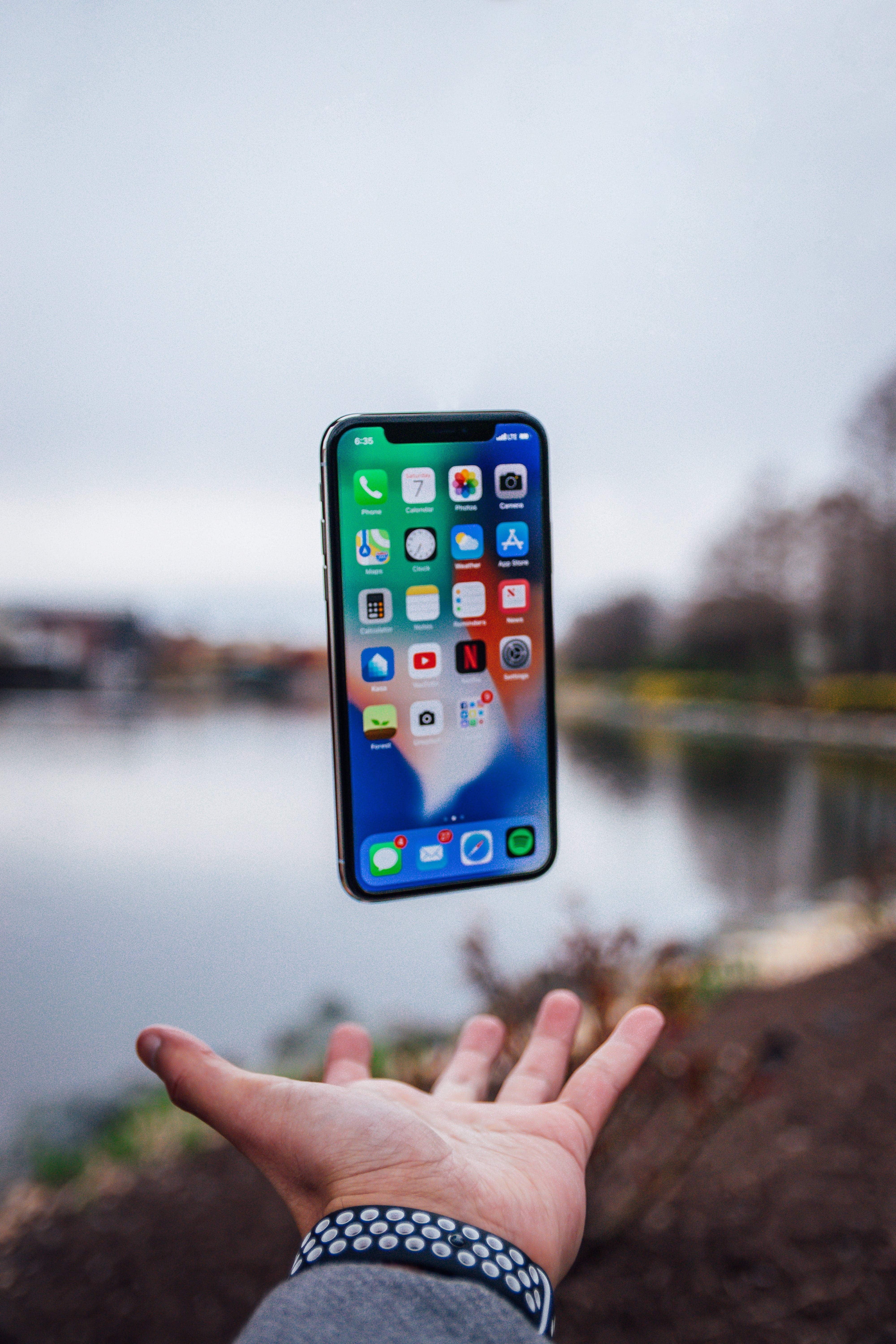 A person's hand is extended upward as an iPhone appears to levitate above it, showcasing various app icons. The background features an outdoor scene with a body of water and trees, indicating a park or riverside location. It's a perfect blend of iOS magic with nature's tranquility.