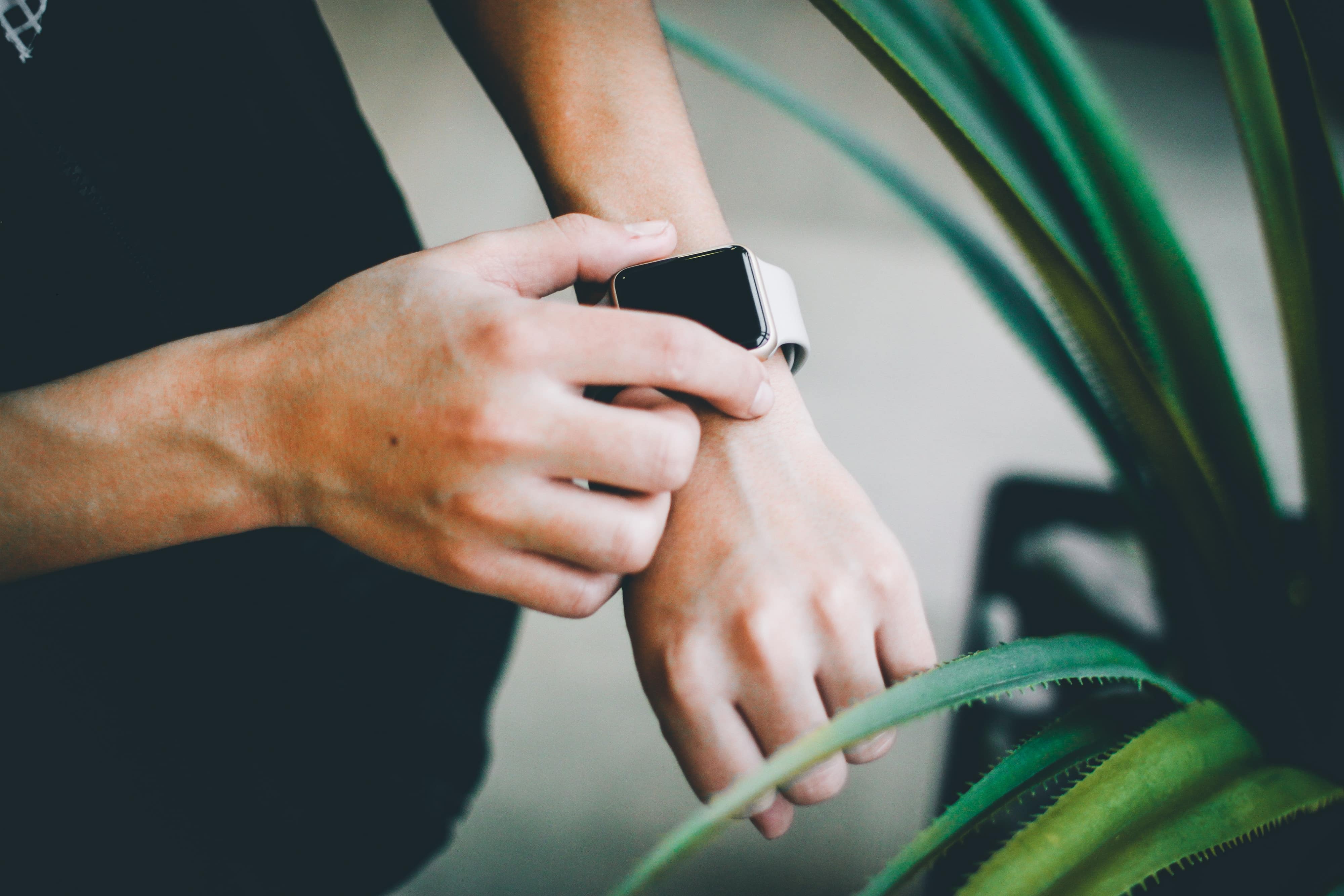 Close-up of a person adjusting their Apple Watch on their wrist. The individual is standing next to some green plants, creating a contrasting background that highlights the smartwatch's ECG functionality.