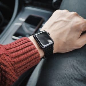 A close-up view of a person's left hand and wrist, adorned with a sleek black Apple Watch. Dressed in a red sweater and gray pants, the individual is inside a car, with parts of the dashboard and a smartphone visible in the background. Did you know one in three prefer an Apple Watch for their smartwatch needs?