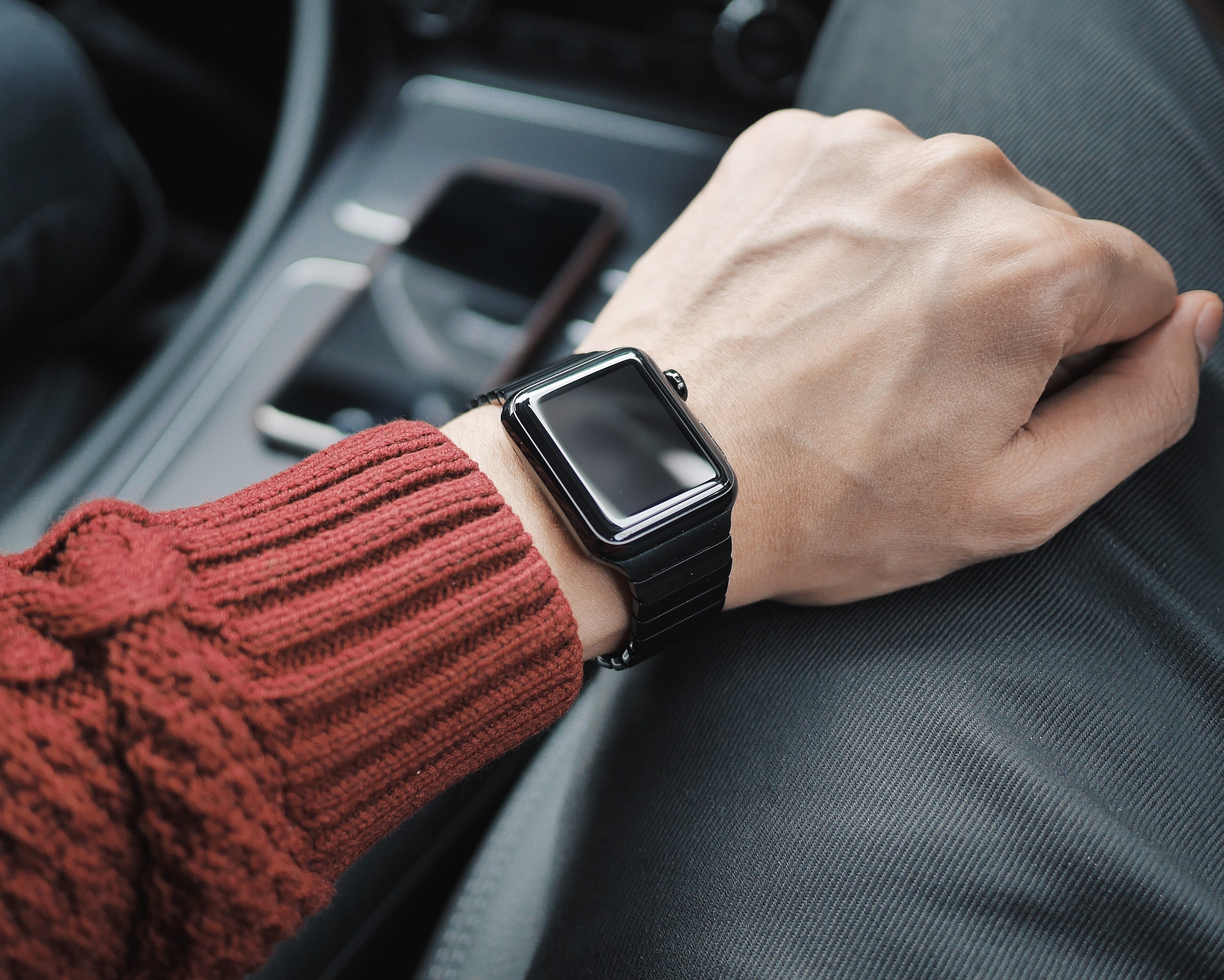 A close-up view of a person's left hand and wrist, adorned with a sleek black Apple Watch. Dressed in a red sweater and gray pants, the individual is inside a car, with parts of the dashboard and a smartphone visible in the background. Did you know one in three prefer an Apple Watch for their smartwatch needs?