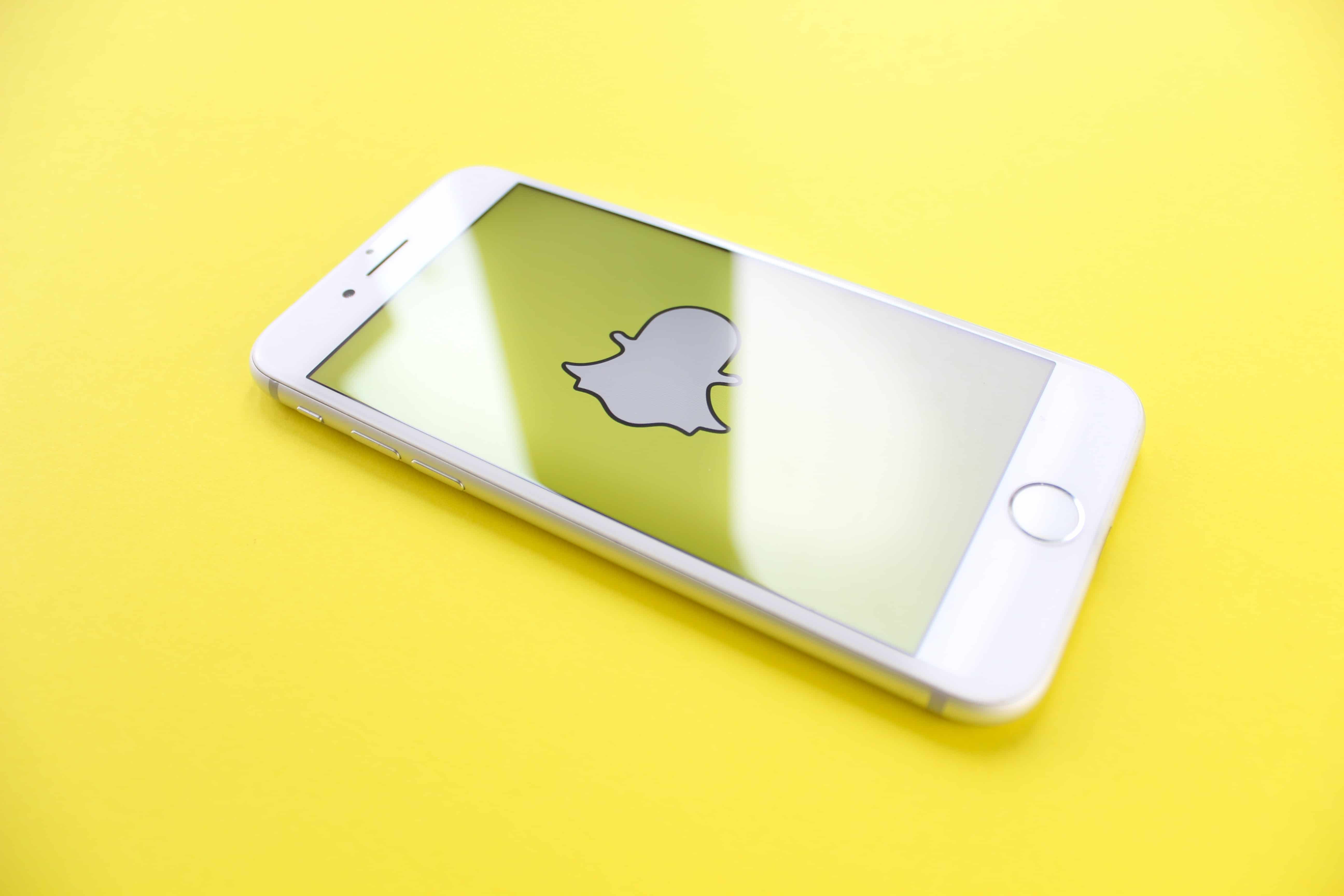 A white smartphone with the Snapchat app open is placed on a yellow surface. The screen displays the Snapchat ghost logo prominently. The phone is angled slightly, reminding users of past concerns about employees snooping on private messages.