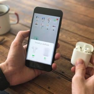 A person holds a pair of Apple AirPods in their right hand and a smartphone displaying the AirPods connection status in their left hand. A white mug with a red "X," a brown pencil, and an Apple smartwatch on the person's wrist are visible on a wooden table.