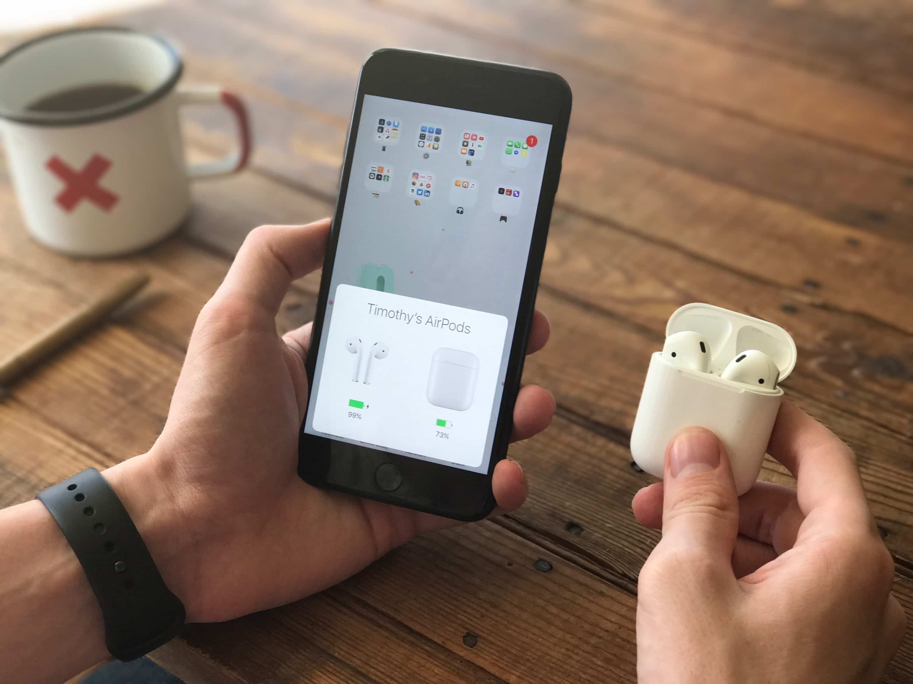 A person holds a pair of Apple AirPods in their right hand and a smartphone displaying the AirPods connection status in their left hand. A white mug with a red "X," a brown pencil, and an Apple smartwatch on the person's wrist are visible on a wooden table.