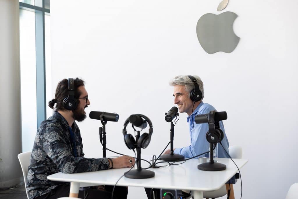 Two men, both wearing headphones and seated at a small table, are engaged in conversation in a podcast setup with microphones and headphones. The background features a large Apple logo, hinting at an iPadOS interview possibly including Craig Federighi.