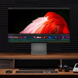 A professional editing suite featuring a large monitor displaying a vivid image of a red and pink bird, flanked by two speakers. The wooden desk holds various editing equipment, including a graphics tablet, keyboard, mouse, and headphones. Apple announces the new Mac Pro as the centerpiece of this setup.