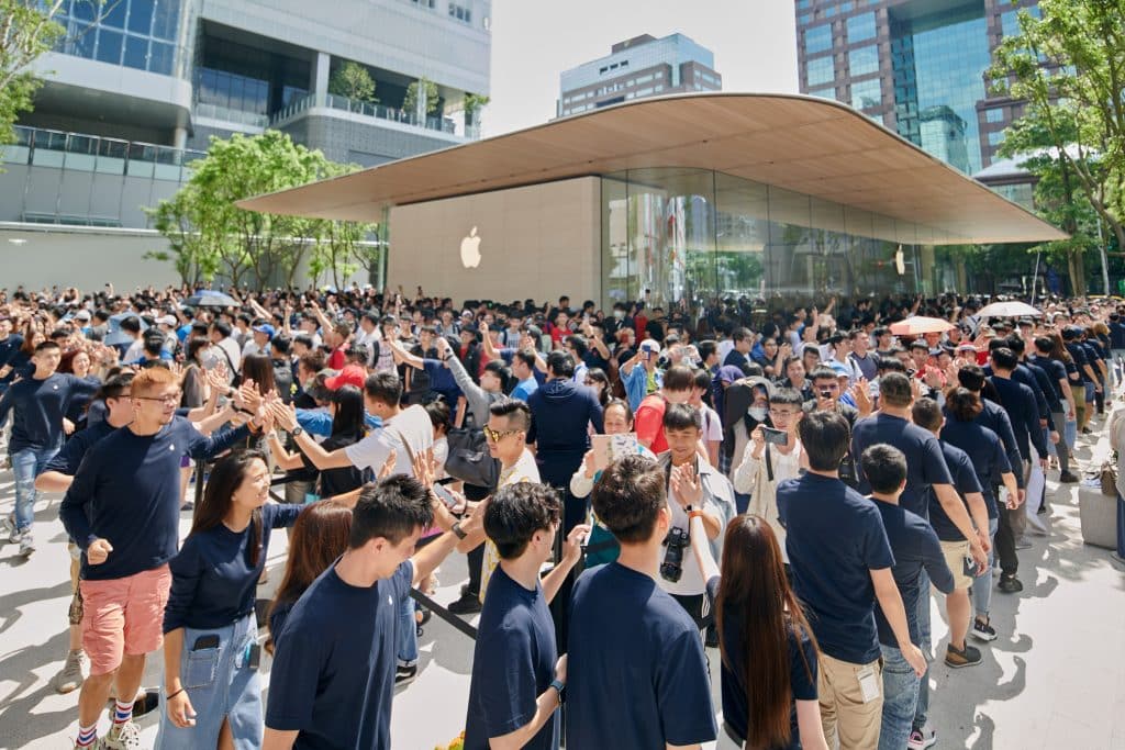 A large crowd gathers outside the latest Apple Store, Apple Xinyi A13, with many holding up phones to take pictures. The store has a modern glass and steel facade. Apple Store employees in navy blue shirts are lined up, forming a pathway and high-fiving visitors.