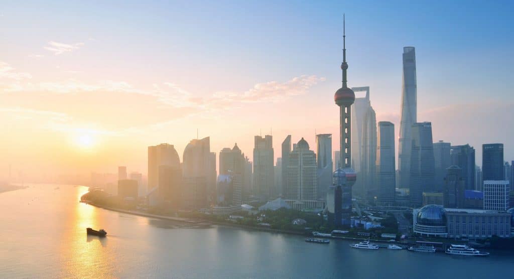 A stunning view of Shanghai's skyline at sunrise, featuring modern skyscrapers including the iconic Oriental Pearl Tower and Shanghai Tower. The cityscape is reflected in the calm waters of the Huangpu River, with a soft golden hue cast over the scene—a subtle reminder of the superpowers' silent competition.