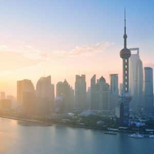 A stunning view of Shanghai's skyline at sunrise, featuring modern skyscrapers including the iconic Oriental Pearl Tower and Shanghai Tower. The cityscape is reflected in the calm waters of the Huangpu River, with a soft golden hue cast over the scene—a subtle reminder of the superpowers' silent competition.