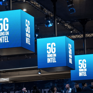 Three blue cube-shaped signs suspended from a ceiling at an event all display the text, "5G RUNS ON INTEL." Below, people are gathered and conversing about exciting advancements like the Intel smartphone modem. Event lighting and large screens in the background amplify the vibrant atmosphere.