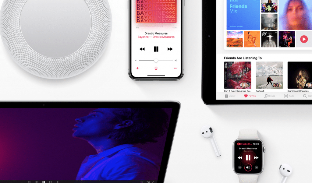 A flat lay of Apple devices including an iPad, iPhone, Apple Watch, MacBook, HomePod, and a pair of AirPods. The iPad and iPhone display music apps while the Apple Watch shows a "Now Playing" screen. This scene highlights the connected Apple ecosystem and its seamless access to Apple Music's 60 million paying subscribers.