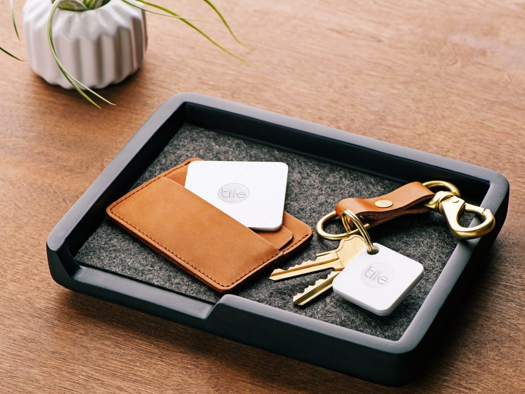 A black tray holds a brown leather wallet with a Tile tracker partially visible inside, next to a keychain with two keys and another Apple Tag attached. A white ceramic vase with a small plant is placed near the tray on a wooden surface.