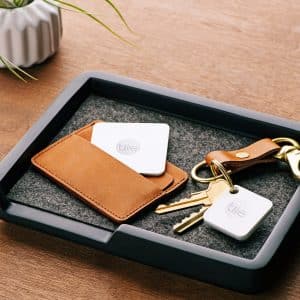 A black tray holds a brown leather wallet with a Tile tracker partially visible inside, next to a keychain with two keys and another Apple Tag attached. A white ceramic vase with a small plant is placed near the tray on a wooden surface.