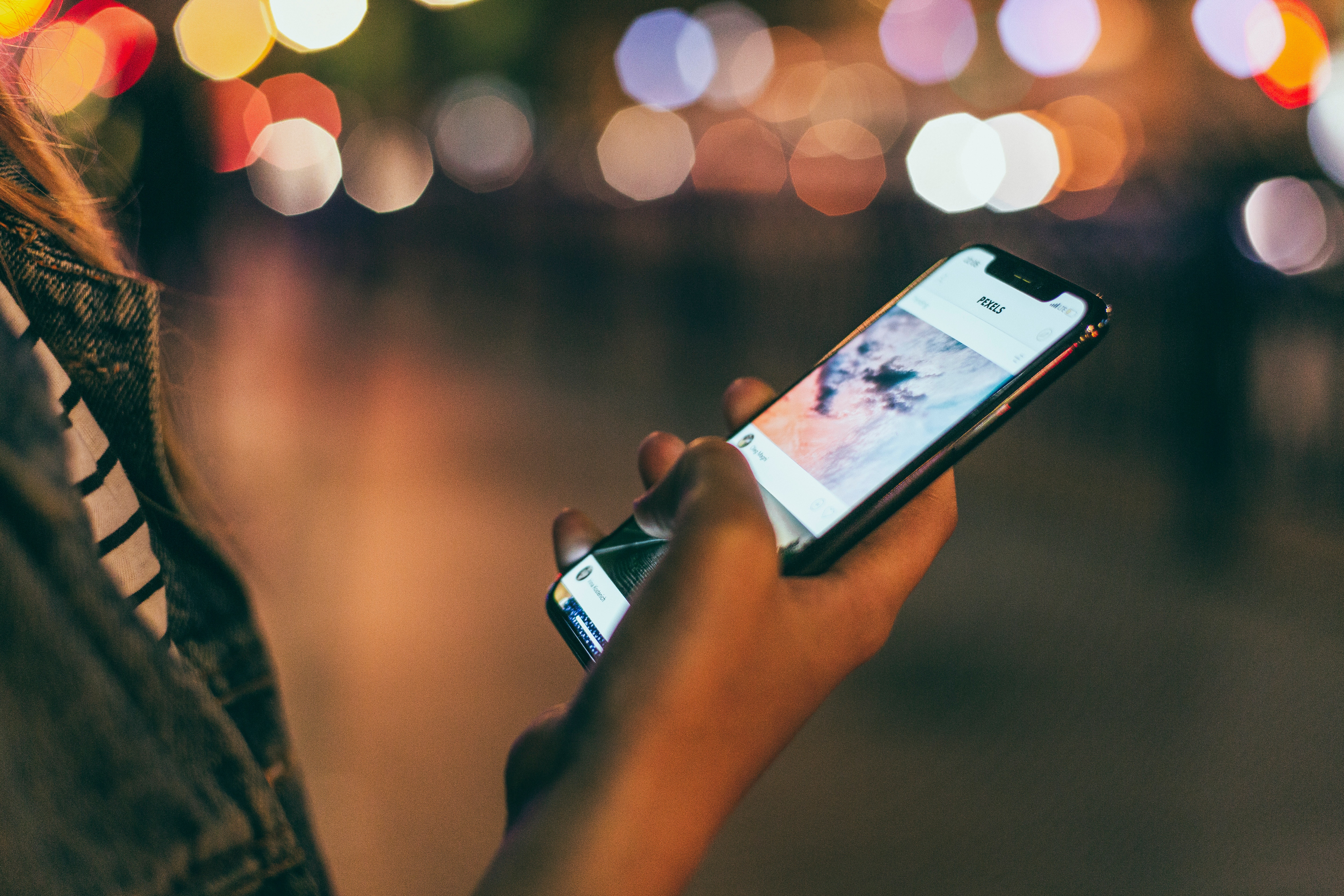 A person is using a smartphone outdoors at night, illuminated by colorful bokeh lights in the blurred background. The phone screen displays an app with multiple images or posts. Part of a denim jacket is visible as they browse casually, perhaps enjoying Apple's latest launch featuring a powerful 5G modem.