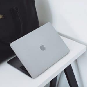 A sleek, partially open silver MacBook Pro sits on a white desk with black legs. A large black leather bag with handles is positioned next to the Apple laptop, creating a contrast against the minimalist, clean background.