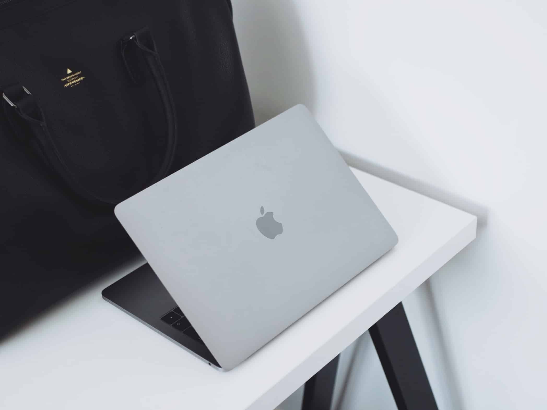 A sleek, partially open silver MacBook Pro sits on a white desk with black legs. A large black leather bag with handles is positioned next to the Apple laptop, creating a contrast against the minimalist, clean background.