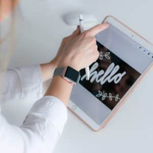 A person with a smartwatch on their left wrist is using a stylus on a tablet running iOS 13. The tablet screen displays the word "hello" in cursive with decorative elements. The person is wearing a white shirt and is seated at a white desk with a computer mouse nearby.