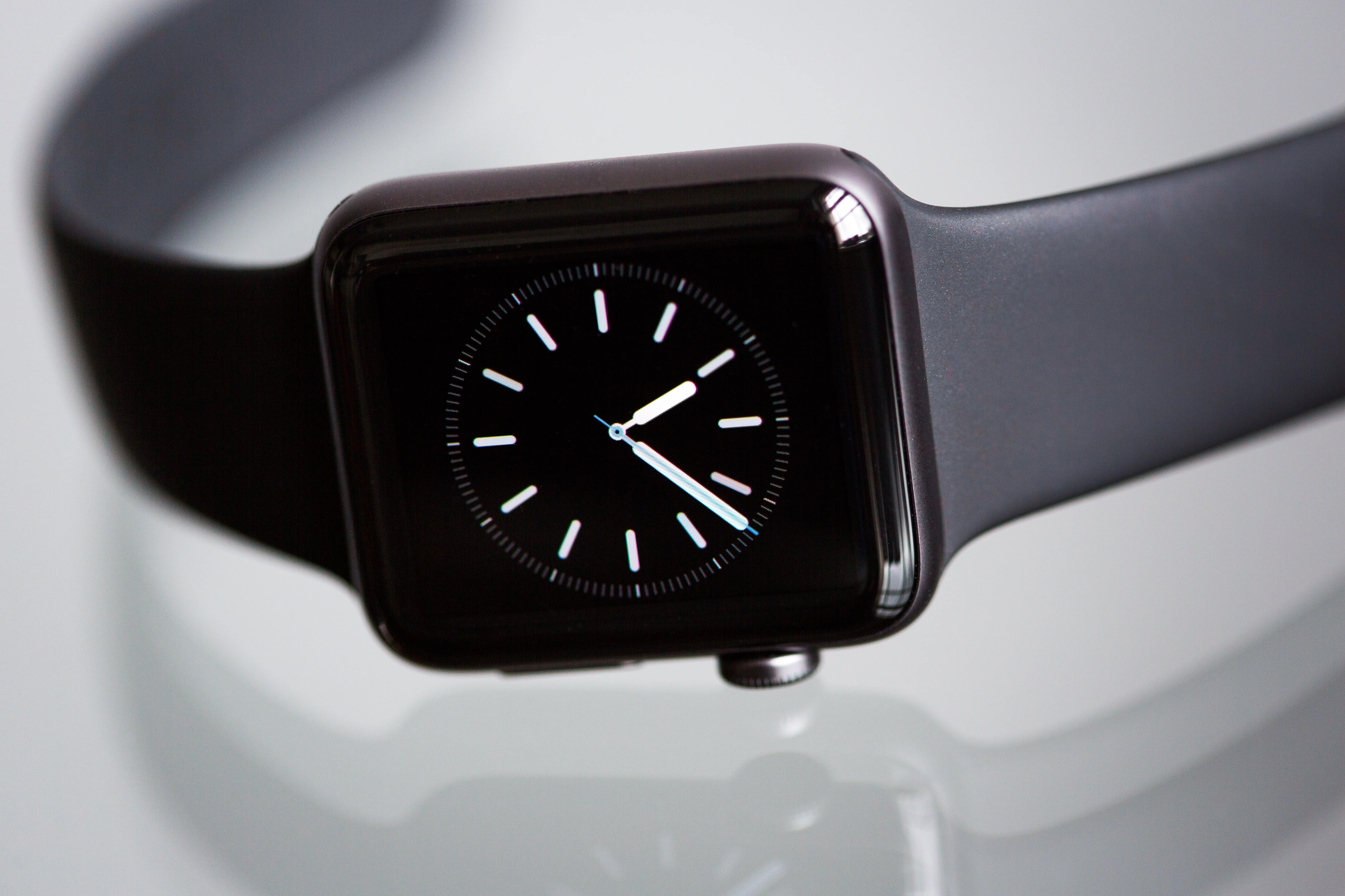 A black Apple smartwatch with a rectangular screen displaying an analog clock face with white hands and tick marks. The watch is on a gray surface, and the black band is visible, showcasing the elegance of watchOS updates received over-the-air.