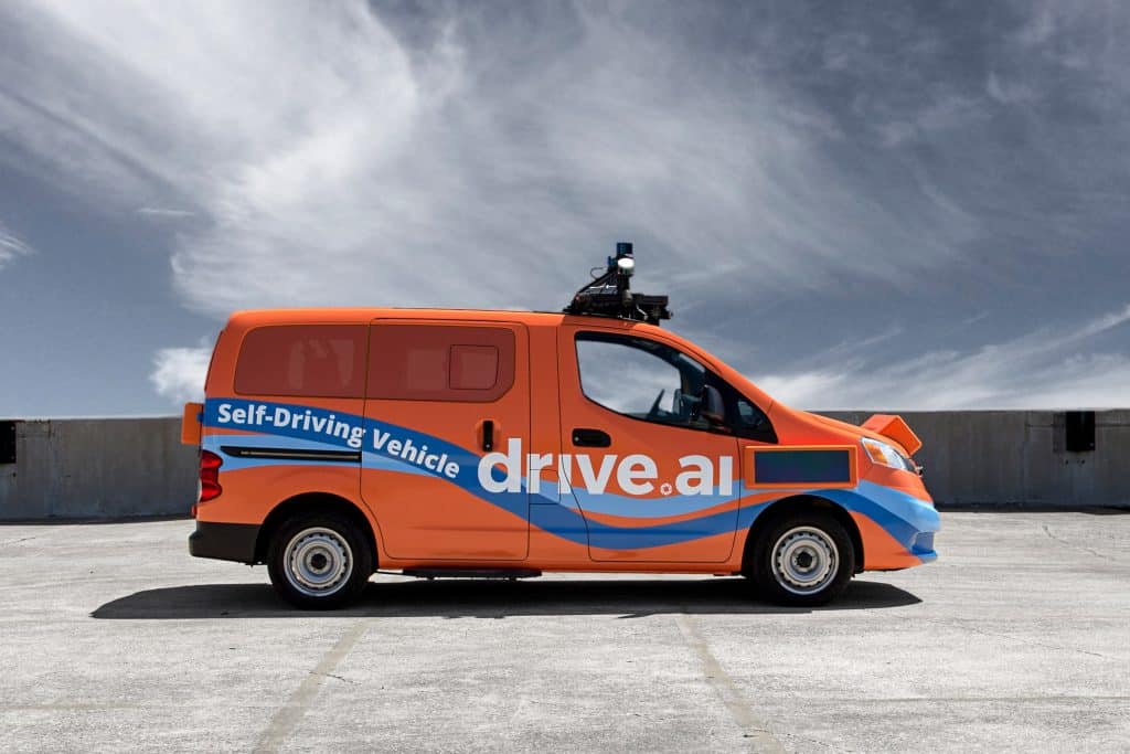 A bright orange van with "Self-Driving Vehicle" and "Drive.ai" written on the sides is parked on a concrete surface under a blue sky with wispy clouds. The van, part of an autonomous vehicle program, is equipped with sensors and cameras on the roof for self-driving capabilities.