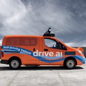 A bright orange van with "Self-Driving Vehicle" and "Drive.ai" written on the sides is parked on a concrete surface under a blue sky with wispy clouds. The van, part of an autonomous vehicle program, is equipped with sensors and cameras on the roof for self-driving capabilities.