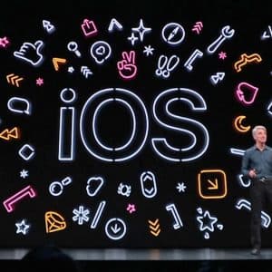 A person stands on a stage with a large display behind them, featuring the text "iOS" in the center. Surrounding the text are colorful icons representing various app features such as music, health, and settings. Highlighting Apple's commitment to inclusivity, the stage is set for a tech presentation focused on accessibility.