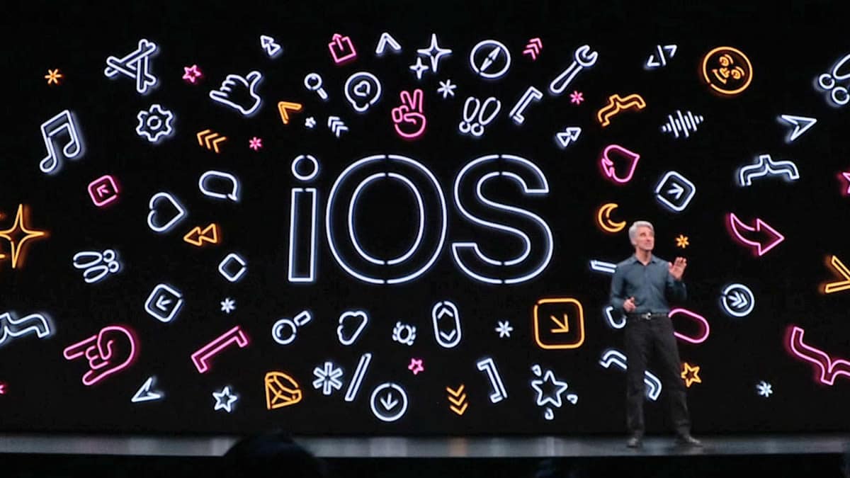 A person stands on a stage with a large display behind them, featuring the text "iOS" in the center. Surrounding the text are colorful icons representing various app features such as music, health, and settings. Highlighting Apple's commitment to inclusivity, the stage is set for a tech presentation focused on accessibility.