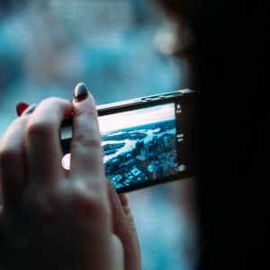 Close-up of a person holding a smartphone horizontally, capturing videos and photos of a cityscape. The background shows blurred buildings and a river, suggesting an elevated view. The person's fingers are partly visible, with nails painted in dark colors.