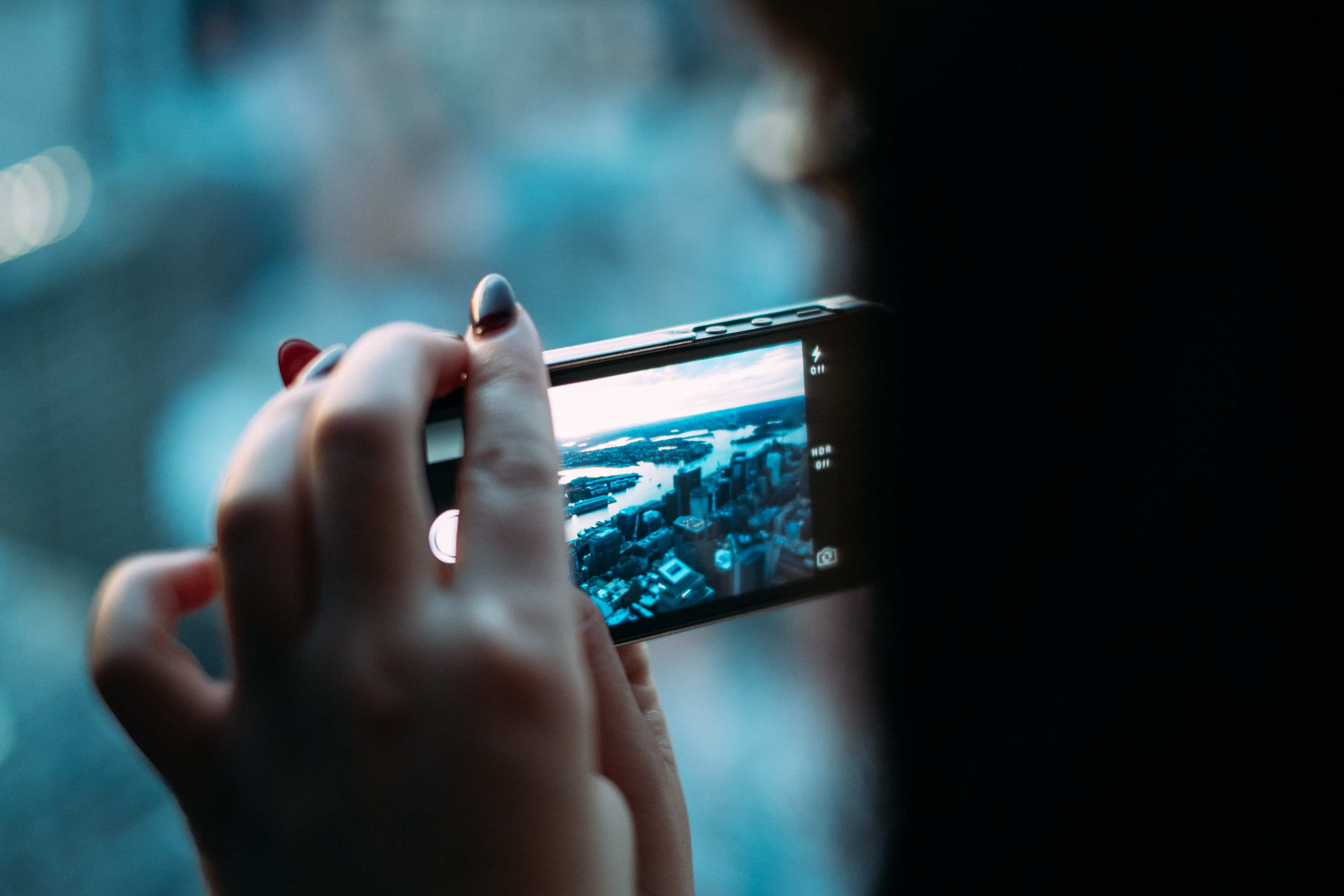 Close-up of a person holding a smartphone horizontally, capturing videos and photos of a cityscape. The background shows blurred buildings and a river, suggesting an elevated view. The person's fingers are partly visible, with nails painted in dark colors.