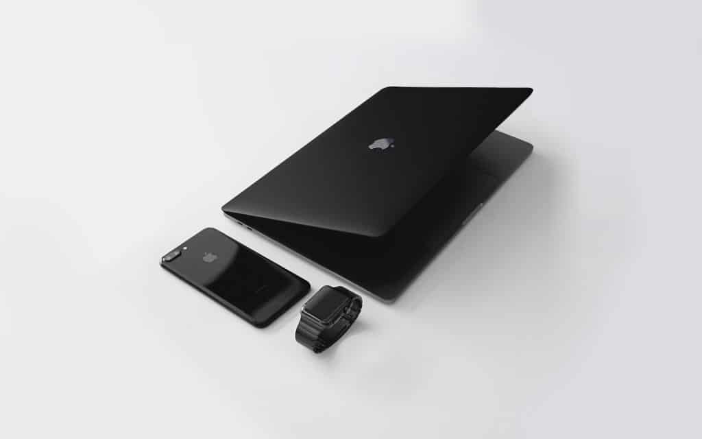 A sleek, black 16-inch MacBook Pro, partially closed with an Apple logo on the lid, sits next to a matching black smartphone and a black smartwatch with a metal strap. They are all arranged on a clean, white surface, showcasing Apple's stunning design aesthetics in preparation for September's new releases.