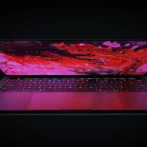 A sleek, partially open Apple laptop with an ARM Mac design features a dark keyboard and a display screen showcasing vibrant red and pink abstract colors. The device is set against a black background, enhancing the vividness of the display.